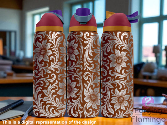 Three brown Owala tumblers featuring a laser-engraved leather tooling in a western floral design with large flowers and swirling paisley-style scrollwork. The wraparound pattern is engraved in white and highly detailed. Each tumbler has a rose-colored lid with a gold accent band and differently colored spouts. The background shows a softly lit indoor setting with desks and windows. Forging Flamingo logo in the bottom right.