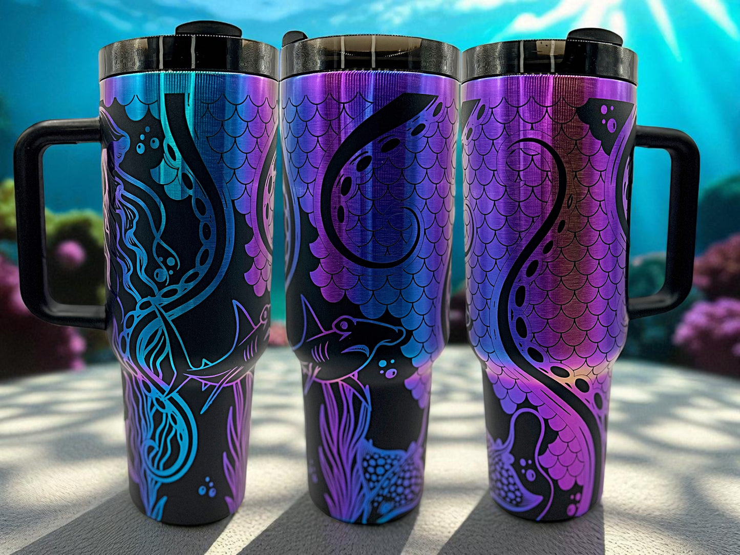 Three iridescent gradient tumblers with handles, shifting from aqua blue to magenta and purple tones. The tumblers feature a seamless underwater scene engraved in black, including a mermaid, shark, jellyfish, and swirling tentacles from a giant octopus. The background is filled with mermaid-scale patterns, coral, seaweed, and bubbles. Set against an oceanic backdrop with sunbeams filtering through water and coral in the distance.