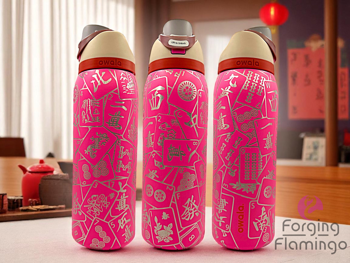 Three bright pink Owala tumblers with laser-engraved mahjong tile designs in white, displayed in a warm, traditional-style room with Chinese decor, including lanterns, calligraphy, and a red tea set. Forging Flamingo logo is visible in the bottom right corner.