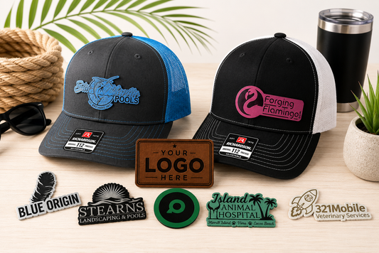 Two customizable trucker hats displayed on a wooden table with lifestyle props like a palm leaf, sunglasses, a tumbler, and a small plant. The left hat is black and blue with a blue fish logo, and the right hat is black and white with a pink flamingo logo. There are a variety of hat patch examples spread out on the table in front of the hats with one patch in the middle with the words "Your Logo Here" engraved on the patch.