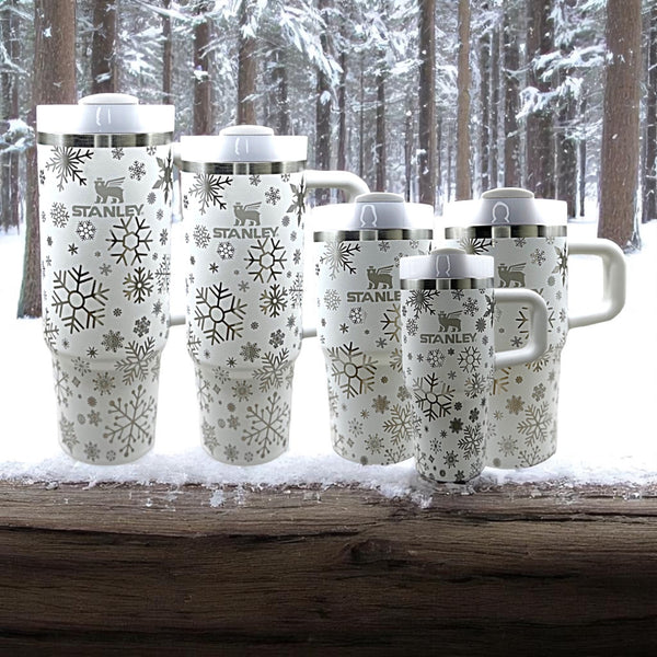 Group of laser-engraved Stanley tumblers in various sizes, featuring a snowflake pattern on a white background, displayed on a snowy log with a winter forest in the background.