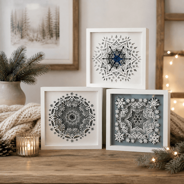 Three framed square wall art pieces with intricate snowflake and geometric mandala cutouts, styled on a wooden shelf with a candle, knit blanket, pine branches, and warm string lights in a cozy indoor setting.
