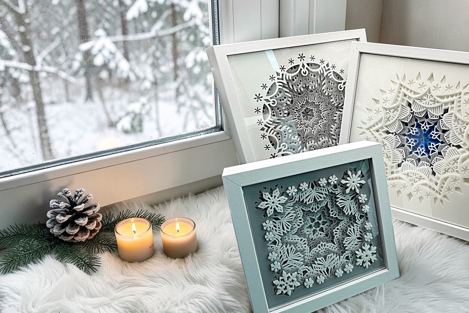 Three framed snowflake art pieces on a windowsill with candles and pine branches.