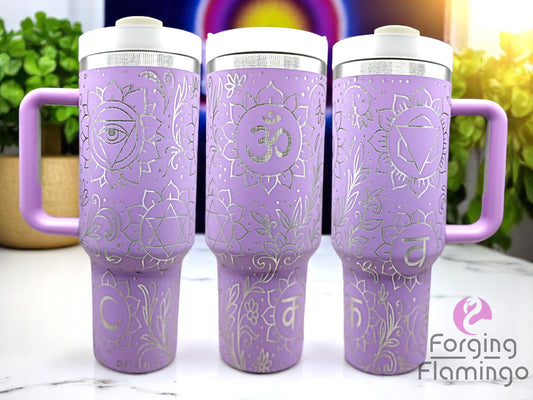 Three lavender handled tumblers with white lids displayed on a white marble surface, featuring an engraved chakra symbol design with intricate floral and swirl patterns. The tumblers have a bright metallic silver engraving, and the background includes green plants and a glowing rainbow light. The Forging Flamingo logo appears in the lower right corner.
