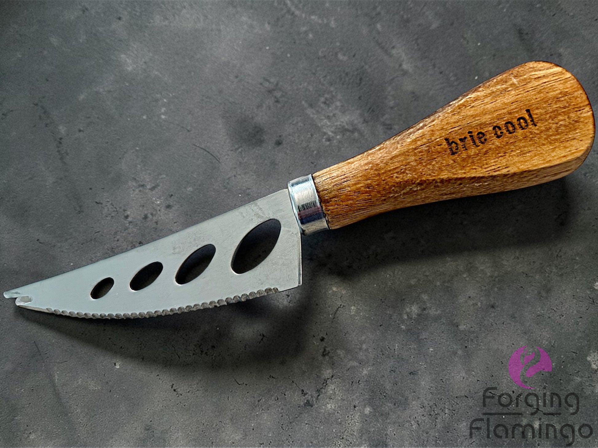 Cheese knife with wooden handle engraved saying brie cool on a dark surface, branded 'Forging Flamingo'.