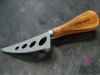 Cheese knife with wooden handle engraved saying brie cool on a dark surface, branded 'Forging Flamingo'.