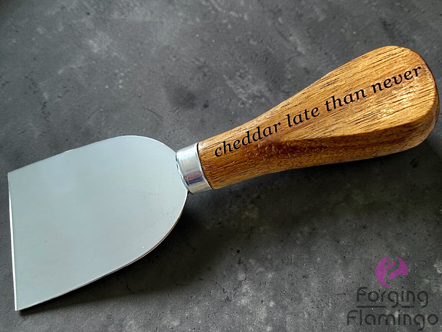 Cheese knife with wooden handle and engraved text saying cheddar late than never on a gray surface, branded 'Forging Flamingo'.
