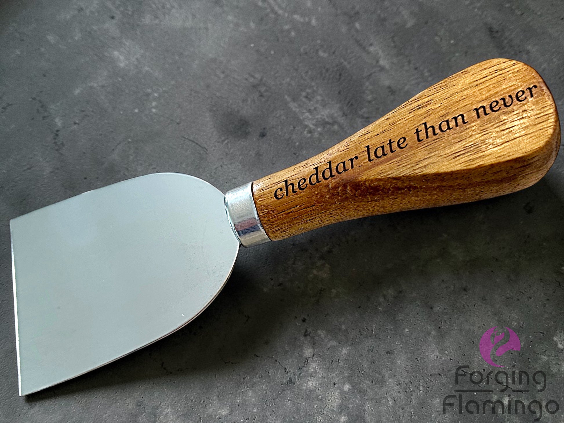 Cheese knife with wooden handle and engraved text saying cheddar late than never on a gray surface, branded 'Forging Flamingo'.