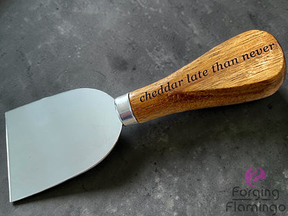 Cheese knife with wooden handle and engraved text saying cheddar late than never on a gray surface, branded 'Forging Flamingo'.