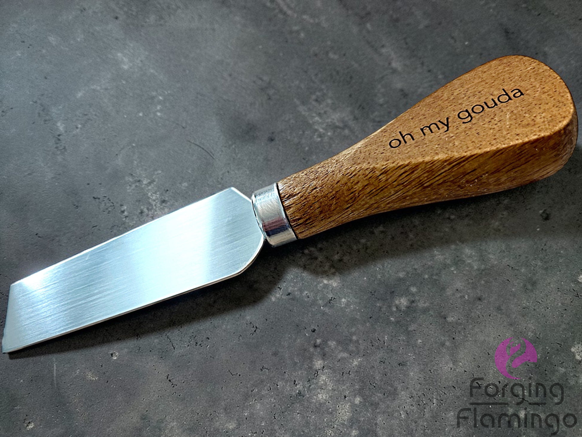 Cheese knife with wooden handle and 'oh my gouda' engraving on a gray surface, brand 'Forging Flamingo' visible.
