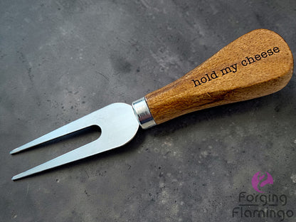 Wooden-handled fork with 'hold my cheese' text on a gray surface, branded 'Forging Flamingo'.