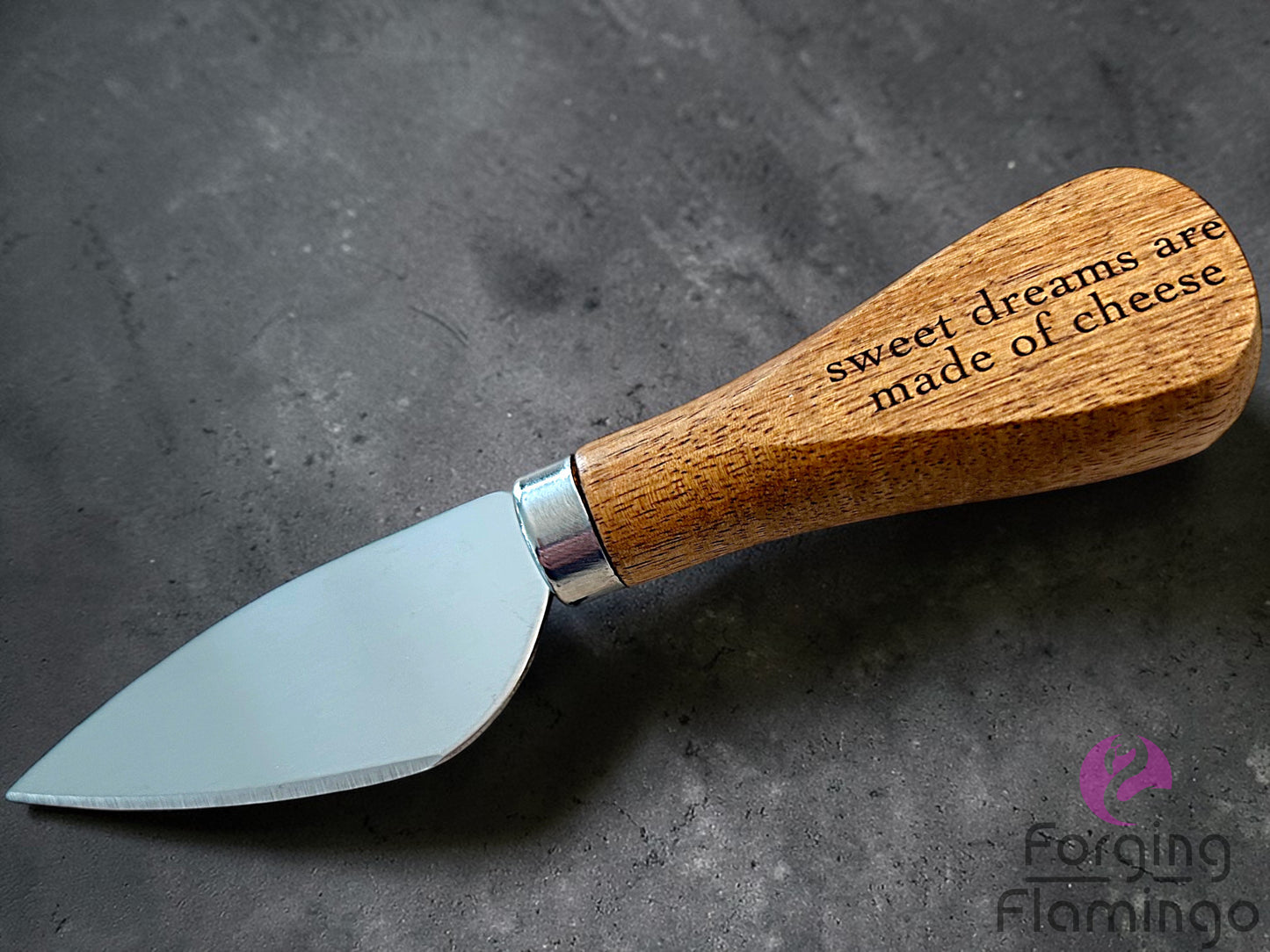 Knife with wooden handle and engraved text saying sweet dreams are made with cheese on a dark surface, brand 'Forging Flamingo' visible.