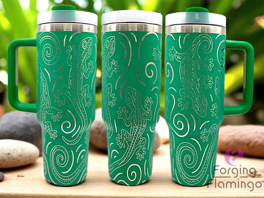 Three green tumblers with laser-engraved geckos and swirling line patterns, displayed on a wooden surface with smooth stones and lush green plants in the background. Forging Flamingo logo is visible in the bottom right corner.
