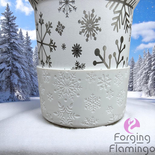 Close-up of a Forging Flamingo tumbler featuring engraved snowflakes above a white silicone boot with embossed snowflake patterns, set against a snowy winter forest backdrop.