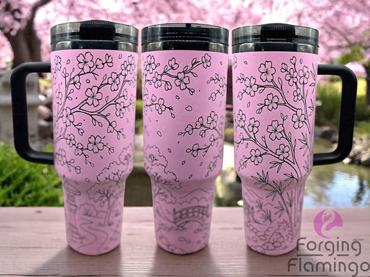 Three pink tumblers engraved with a seamless Japanese Garden design featuring cherry blossoms, a bridge, and bamboo, displayed on a wooden surface with a tranquil pond and blooming cherry trees in the background.