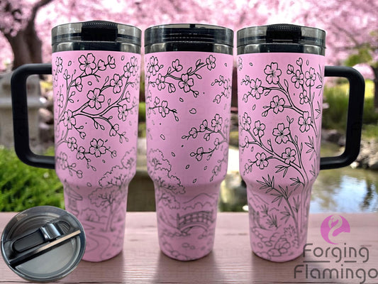 Three pink laser-engraved tumblers with cherry blossom branch designs, displayed outdoors in front of a scenic spring garden with blooming trees and a pond. Inset image shows a close-up of the lid. Forging Flamingo logo is visible in the bottom right corner.
