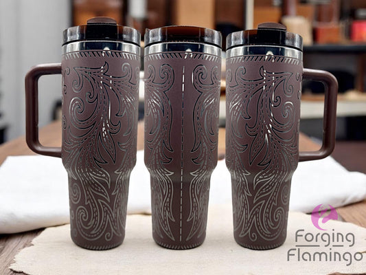 Three brown handled tumblers with clear lids and white straws, displayed on a marble surface. The tumblers feature an engraved Western-inspired tooled leather pattern of stitched swirling leaf motifs, revealing a silver metal base beneath. The design wraps fully around each tumbler. One tumbler is positioned to show a vertical stitched seam detail. A stack of leather pieces sits to the right, and the Forging Flamingo logo is in the lower right corner.
