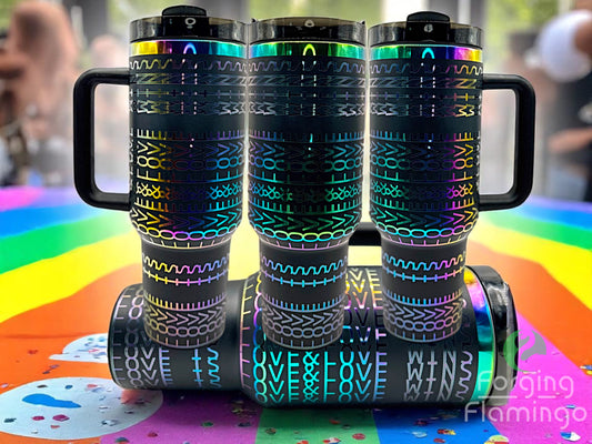 Three black tumblers with rainbow-reflective laser-engraved geometric and ‚ÄúLOVE WINS‚Äù patterns, displayed on a vibrant rainbow table with confetti, celebrating Pride. One tumbler is laid horizontally in front. Forging Flamingo logo is visible in the bottom right corner.
