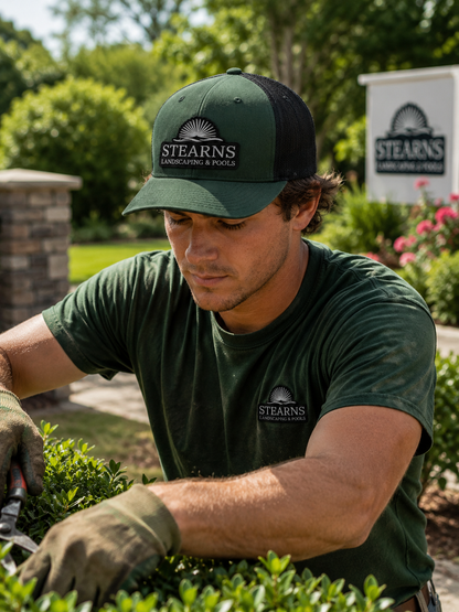 A man wearing a dark green and black Stearns cap and matching Stearns t-shirt is shown gardening outdoors while using pruning shears. The Stearns logo is visible on both the cap and the shirt.